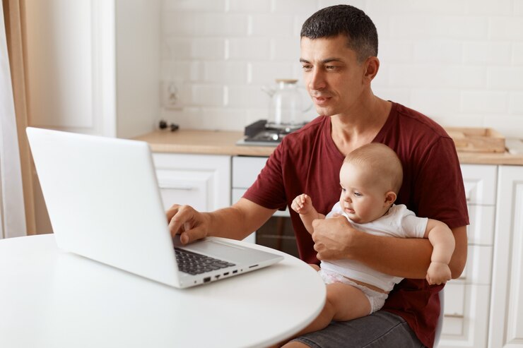 indoor-shot-handsome-man-with-dark-hair-wearing-burgundy-casual-t-shirt-working-laptop-while-babysitting-looking-notebook-screen-posing-white-kitchen_176532-15474